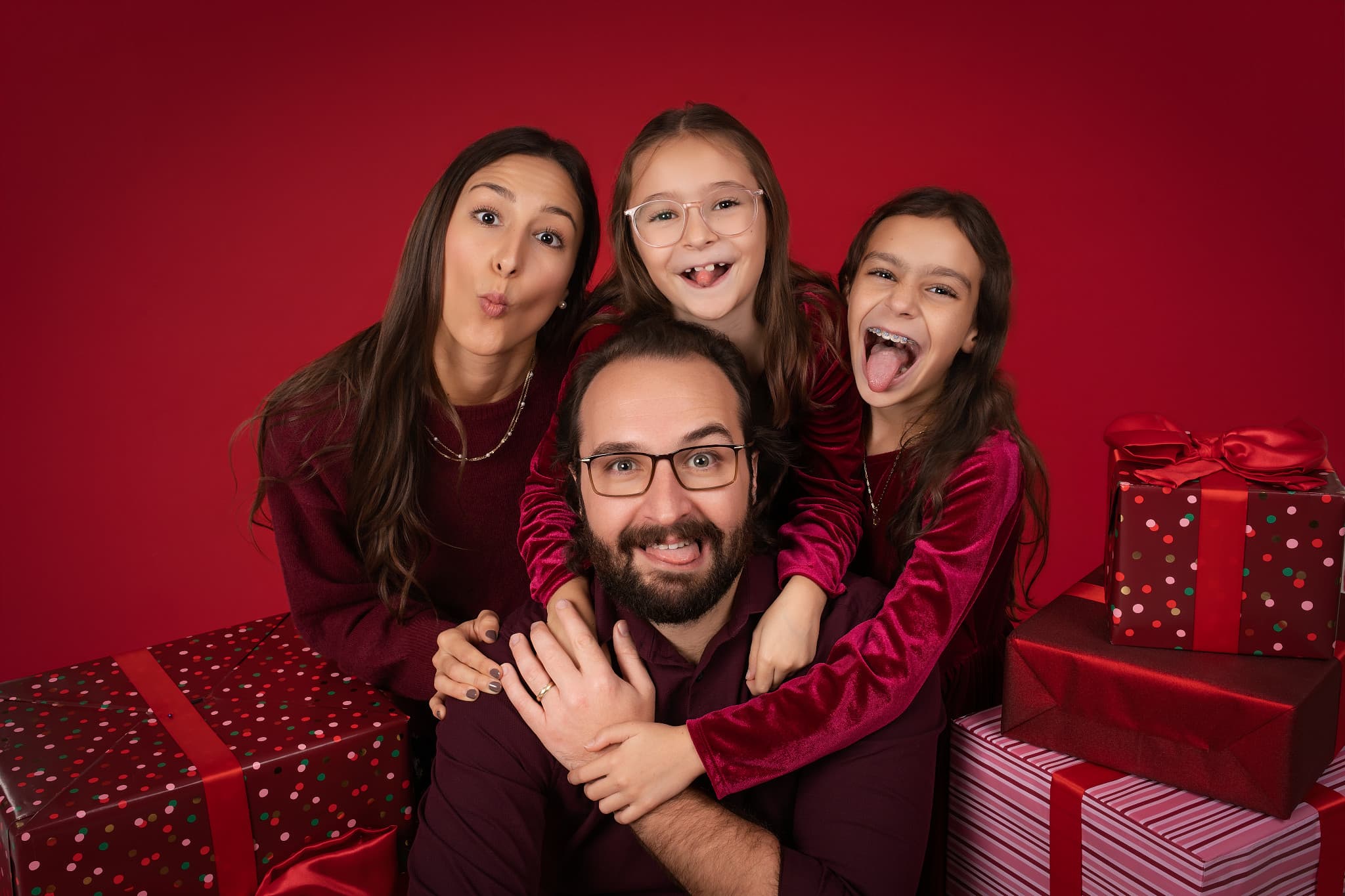 A family of four laughing together against a deep red backdrop, surrounded by wrapped gifts