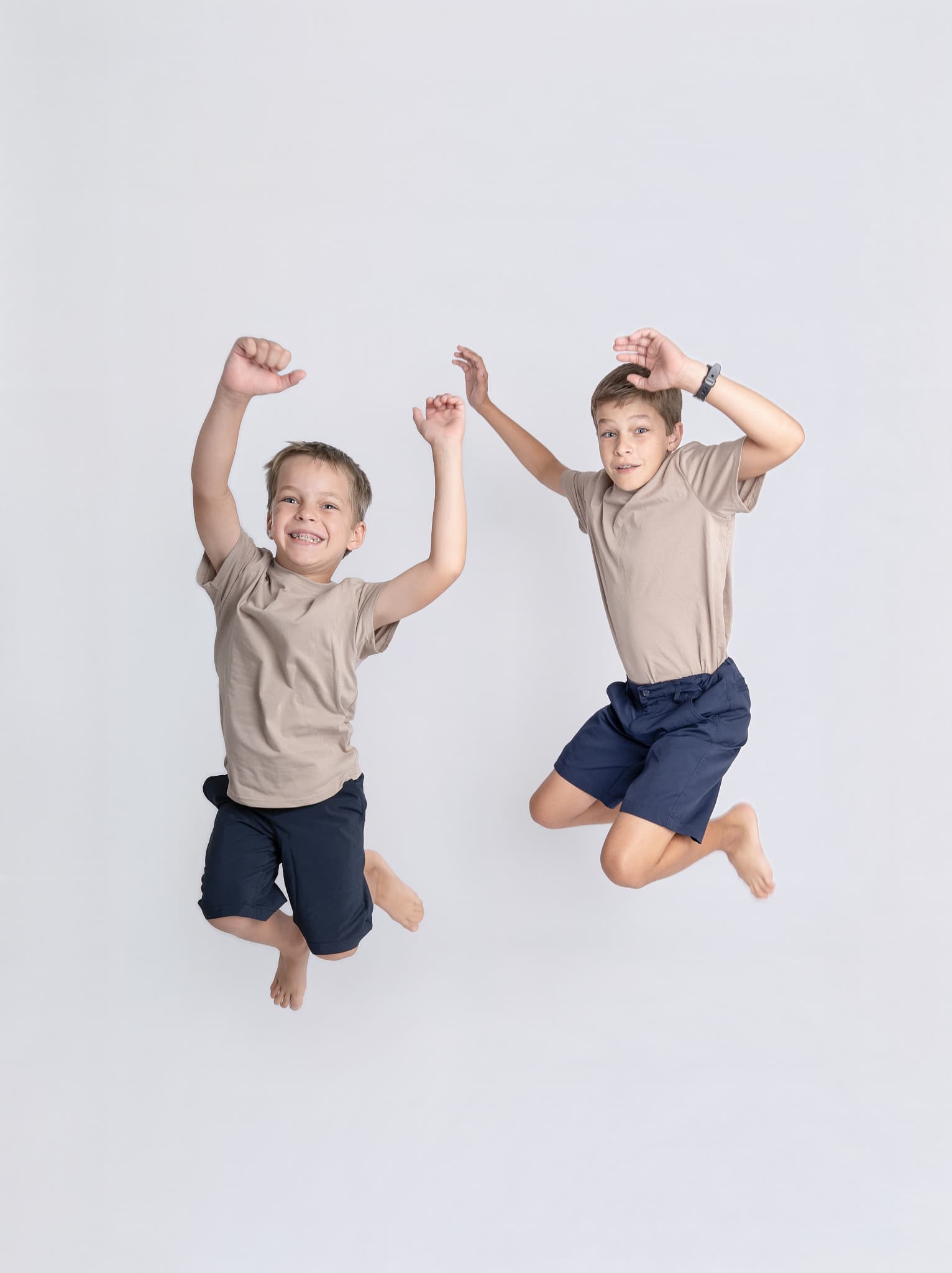 Two brothers mid-jump against a plain studio wall, both barefoot and laughing