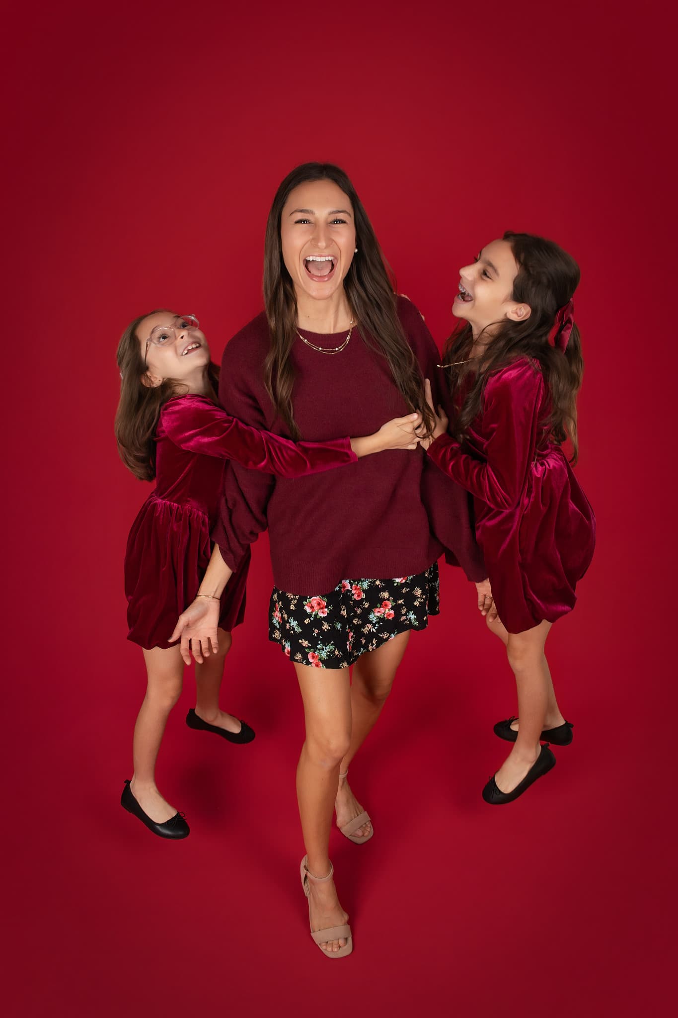 A mother laughing with her two daughters against a deep red backdrop, all three in burgundy velvet