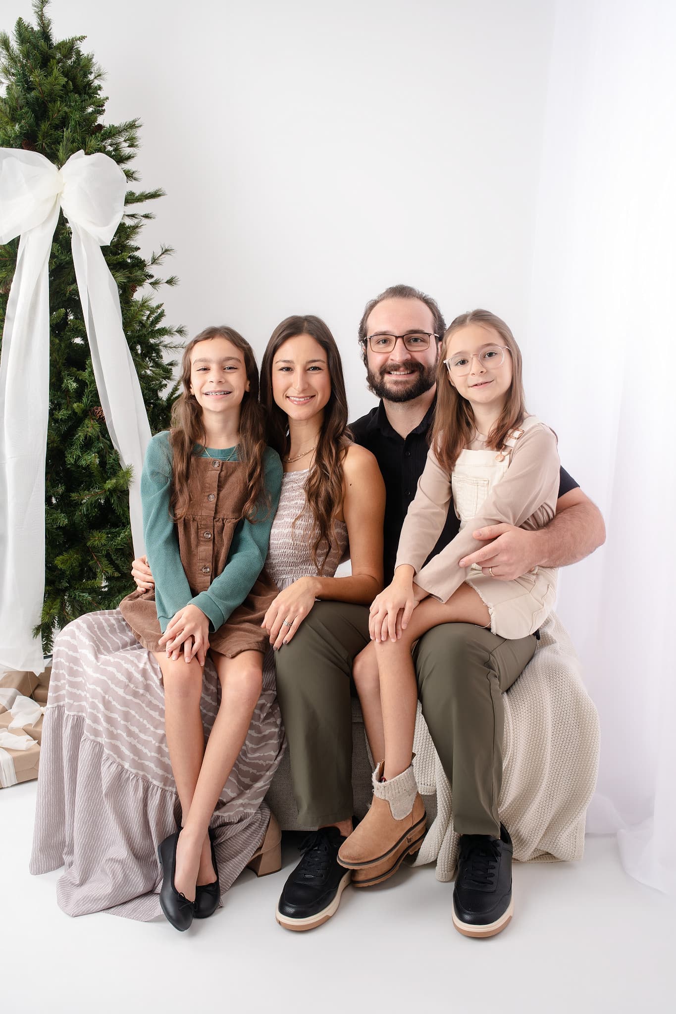 A family of four on a bench beside a white Christmas tree tied with a wide white bow, in the Brandon studio