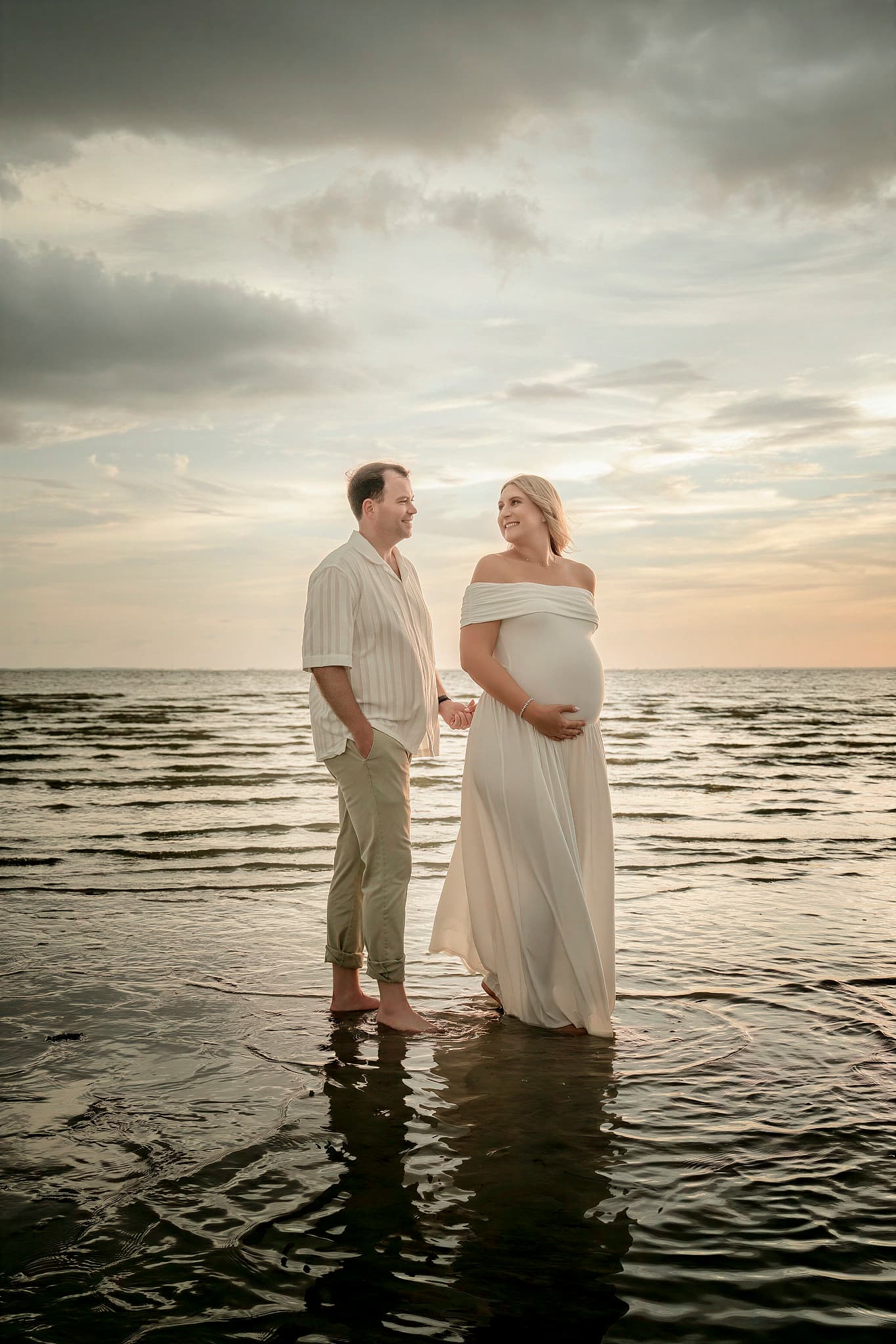 Expectant parents holding hands in shallow water at sunset, the mother in a long white gown