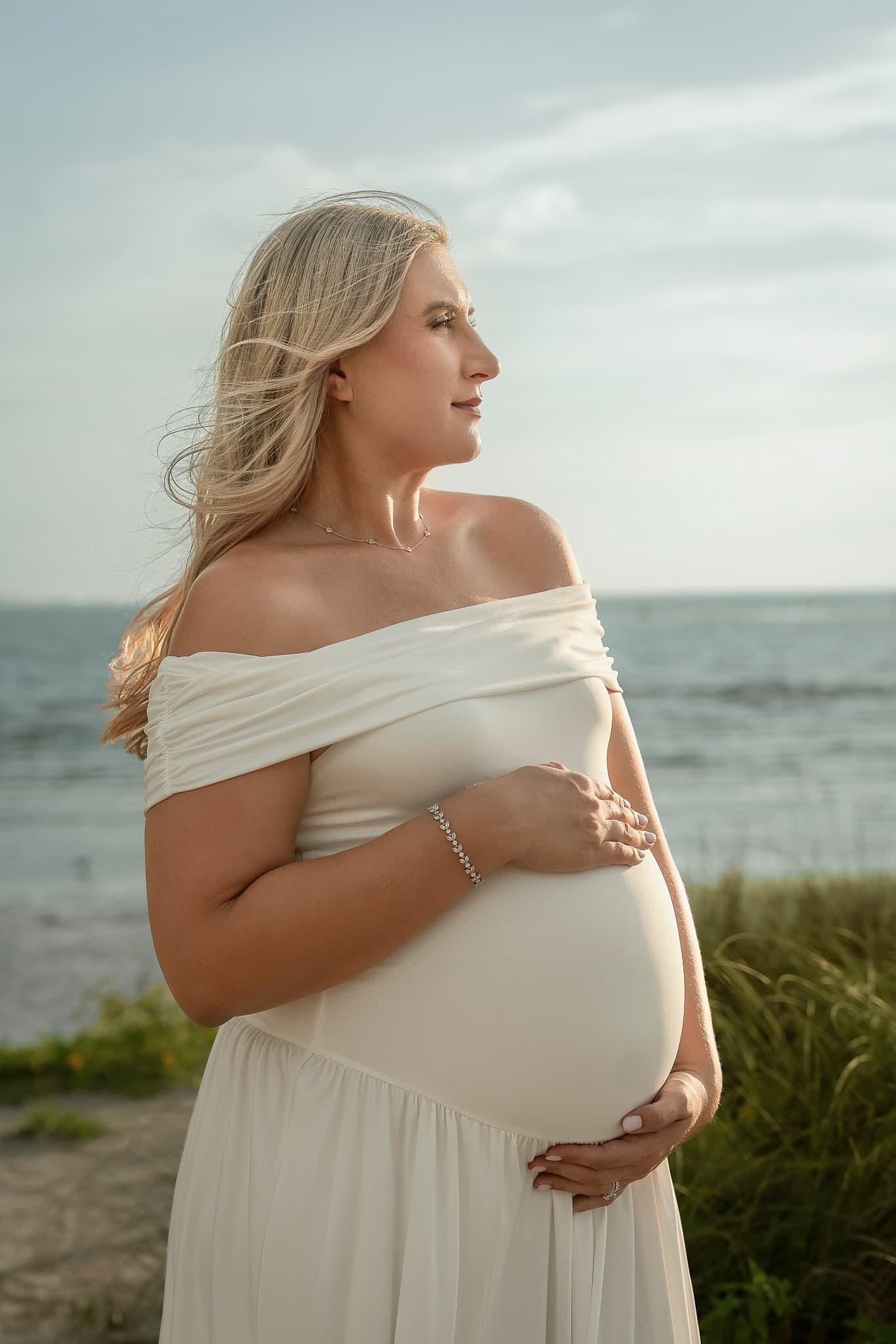 An expectant mother in a cream off-shoulder gown at the shoreline, both hands cradling her belly